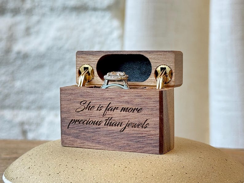K&ouml;nnte beinhalten: Eine rechteckige Ringbox aus Holz mit Klappdeckel und goldfarbenen Metallscharnieren. Die Vorderseite ist mit dem Text "She is far more precious than jewels" graviert. Im Inneren befindet sich ein silberfarbener Ring mit einem zentralen Stein und einer Halo-Fassung.