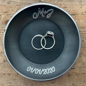 May include: A dark gray metal ring dish with the initials "M+J" and the date "01/01/2020" engraved in white. Two silver rings with clear stones are inside the dish. The dish is on a wooden surface.