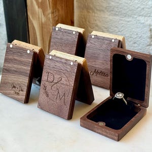 May include: A collection of rectangular dark wood ring boxes with magnetic closures. Some boxes feature laser-engraved text including names and dates. One open box displays a silver ring with a clear stone set against a black velvet interior.
