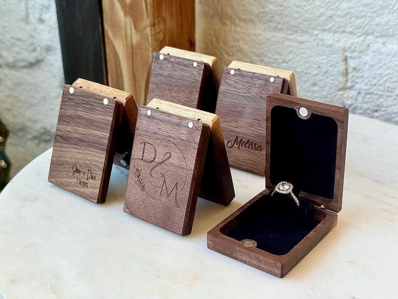 May include: A collection of rectangular dark wood ring boxes with magnetic closures. Some boxes feature laser-engraved text including names and dates. One open box displays a silver ring with a clear stone set against a black velvet interior.