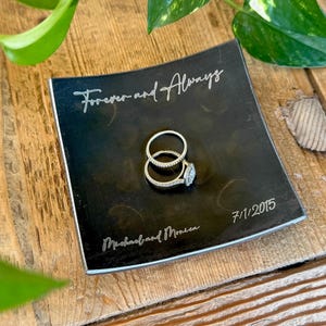 May include: A square black ring dish with curved edges featuring the engraved text Forever and Always, Michael and Monica, and 7/1/2015. Two silver-toned rings, one with a central stone, are resting in the center of the dish.