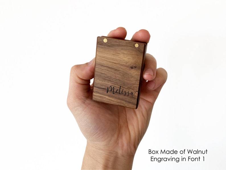 May include: A small wooden box made of walnut with a brass hinge and a personalized inscription that reads "Melissa" in a cursive font. The box is held in a person's hand.