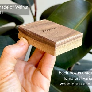 A closed, compact wooden engagement ring box held in hand against a green outdoors background, featuring descriptive text overlays highlighting its solid walnut construction and natural wood grain. Showcasing variation in natural wood grain and color