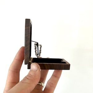 May include: A small wooden box with a hinged lid, open to reveal a gold chain necklace inside. The box is made of dark wood with a natural grain pattern.