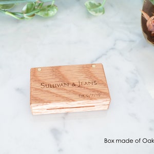 May include: A rectangular light brown oak wood box with a hinged lid featuring two small metal pins. The top of the box is engraved with the text Sullivan & Jeans and Est. 9/7/19 in a dark font. The wood grain is visible across the surface.