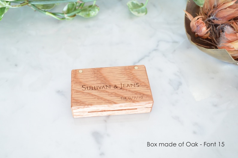 May include: A rectangular light brown oak wood box with a hinged lid featuring two small metal pins. The top of the box is engraved with the text Sullivan & Jeans and Est. 9/7/19 in a dark font. The wood grain is visible across the surface.