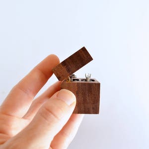 May include: A small wooden ring box with a hinged lid, open and held in a hand. The box is made of dark wood with a natural grain pattern.