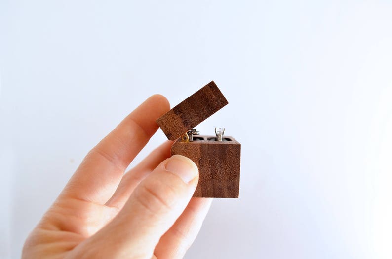 May include: A small wooden ring box with a hinged lid, open and held in a hand. The box is made of dark wood with a natural grain pattern.