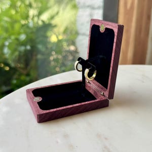 May include: A rectangular purple wooden jewelry box with a hinged lid and a black velvet interior lining. The box is open, displaying a pair of small gold hoop earrings mounted on a central black stand within the velvet-lined compartment.