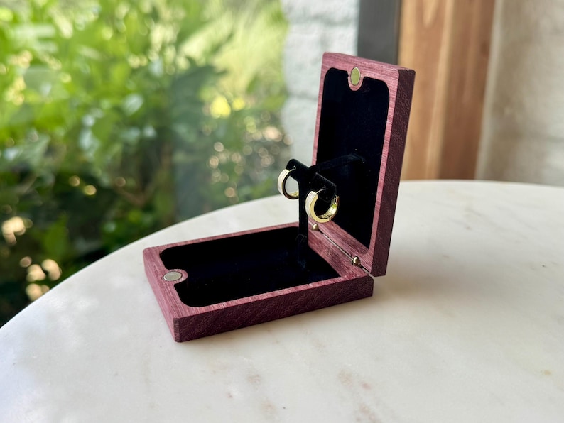 May include: A rectangular purple wooden jewelry box with a hinged lid and a black velvet interior lining. The box is open, displaying a pair of small gold hoop earrings mounted on a central black stand within the velvet-lined compartment.