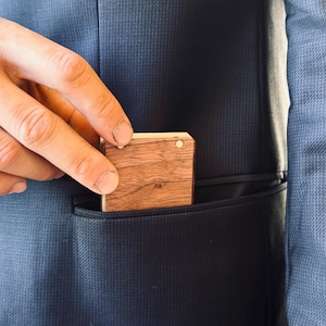 Close-up of a hand pulling a slim, pocket-sized wooden engagement ring box out of a suit jacket pocket, showing the smooth wood grain and compact design.