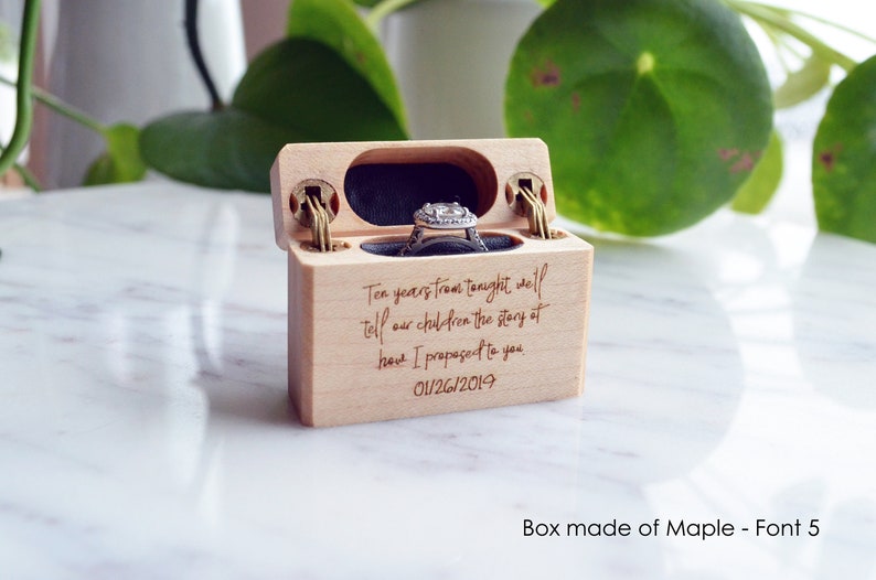 K&ouml;nnte beinhalten: Eine helle Holzringschatulle mit Klappdeckel und dunklem Innenfutter. Auf der Vorderseite ist der Text eingraviert: Ten years from tonight we'll tell our children the story of how I proposed to you 01/26/2019. Ein silberner Ring befindet sich im Inneren.