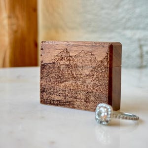 K&ouml;nnte beinhalten: Eine rechteckige Holzschatulle mit einer lasergravierten Berglandschaft auf der Vorderseite, platziert neben einem silberfarbenen Ring mit einem quadratischen Stein in der Mitte und einem mit kleinen klaren Steinen besetzten Ringband.