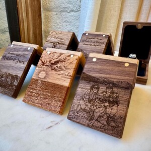 May include: A collection of rectangular wooden ring boxes with hinged lids. Each box features laser-engraved imagery, including mountain landscapes, ocean sunsets, and a couple with a dog. One box is open, revealing a black interior and a ring.