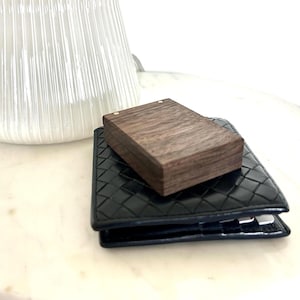 A dark wooden engagement ring box resting open on a dark blue wallet next to a decorative white ceramic ribbed vase, showcasing the small size.