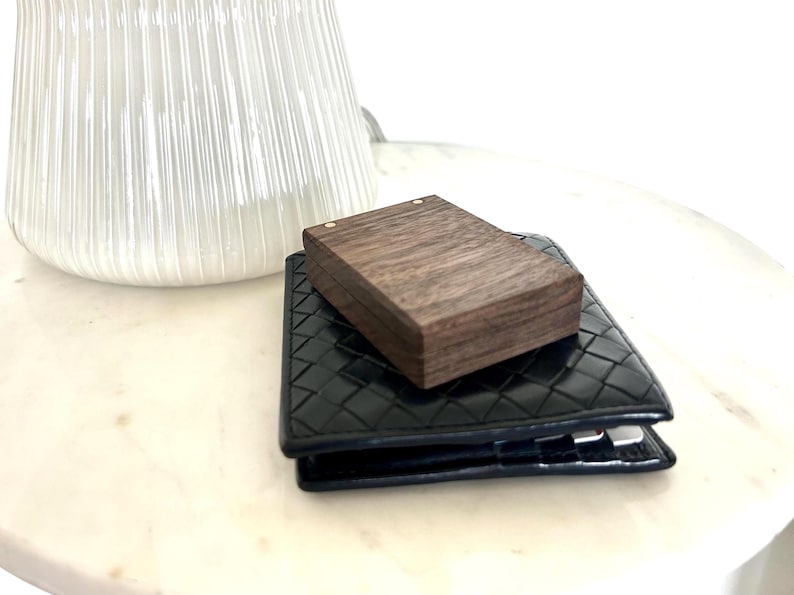 A dark wooden engagement ring box resting open on a dark blue wallet next to a decorative white ceramic ribbed vase, showcasing the small size.