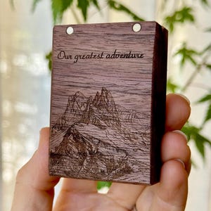 A hand holding a compact wooden engagement ring box featuring a detailed custom line-art engraving of a mountain range and trees, set against a softly blurred green nature background.