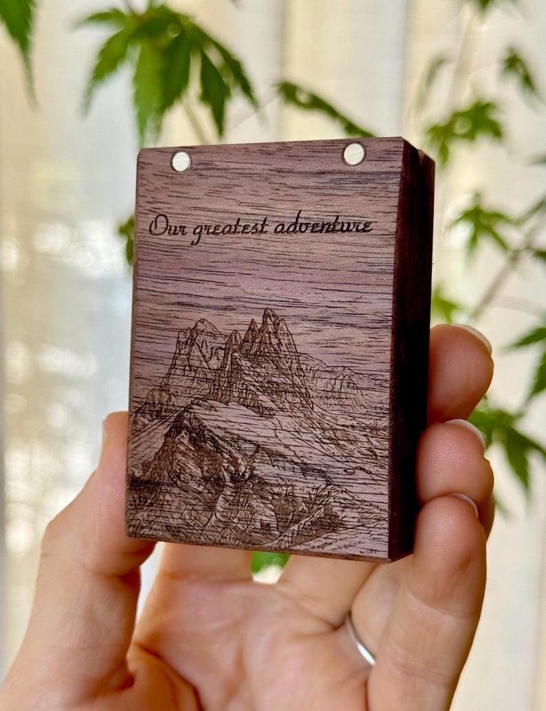 A hand holding a compact wooden engagement ring box featuring a detailed custom line-art engraving of a mountain range and trees, set against a softly blurred green nature background.