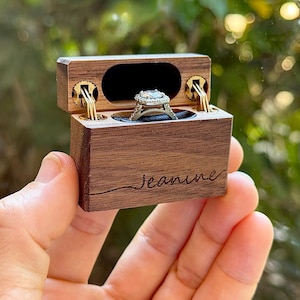 K&ouml;nnte beinhalten: Eine rechteckige Ringbox aus dunklem Holz mit Klappdeckel und goldfarbenen Metallscharnieren. Auf der Vorderseite ist der Name Jeanine in Schreibschrift eingraviert. Im Inneren befindet sich ein silberfarbener Ring mit quadratischer Halo-Fassung.