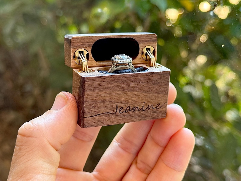 K&ouml;nnte beinhalten: Eine rechteckige Ringbox aus dunklem Holz mit Klappdeckel und goldfarbenen Metallscharnieren. Auf der Vorderseite ist der Name Jeanine in Schreibschrift eingraviert. Im Inneren befindet sich ein silberfarbener Ring mit quadratischer Halo-Fassung.