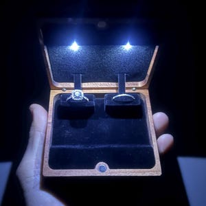 May include: A wooden ring box with a black velvet interior and two compartments. The box is lit from the inside with two white LED lights. A diamond ring is displayed in one compartment and a plain band is displayed in the other compartment.
