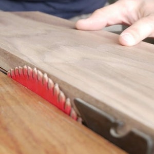 May include: A close-up of a table saw with a piece of wood being cut. The saw blade is red and has a black guard. The wood is light brown and has a smooth surface.
