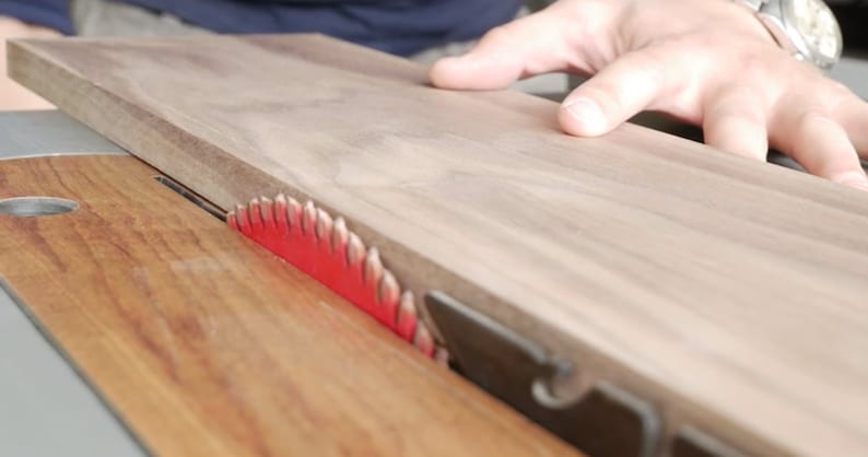 May include: A close-up of a table saw with a piece of wood being cut. The saw blade is red and has a black guard. The wood is light brown and has a smooth surface.