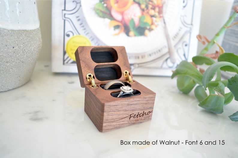 May include: A wooden ring box with two compartments, one for each ring. The box is made of walnut wood and has a gold hinge. The box is engraved with the text "Fetchco" and the date "October 11, 2019".