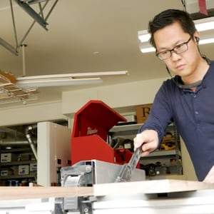 May include: A person wearing glasses and a blue shirt uses a tool to cut a piece of wood on a table saw in a workshop.
