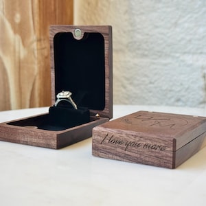 May include: A dark brown wooden ring box with a black velvet interior, holding a silver ring with a square-cut stone. A second closed wooden box sits beside it, featuring the engraved text "I love you more" and the initials "DJM" on the lid.