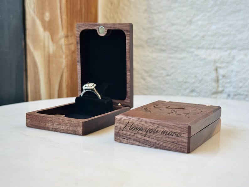 May include: A dark brown wooden ring box with a black velvet interior, holding a silver ring with a square-cut stone. A second closed wooden box sits beside it, featuring the engraved text "I love you more" and the initials "DJM" on the lid.