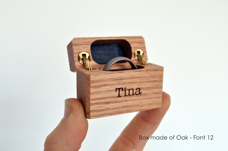 K&ouml;nnte beinhalten: Eine kleine rechteckige Ringbox aus Eichenholz mit Klappdeckel und goldfarbenen Metallscharnieren. Auf der Vorderseite ist der Name Tina eingraviert. Im Inneren befindet sich ein dunkelblauer Samteinsatz, der einen schlichten Metallring h&auml;lt.
