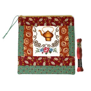 May include: A quilted pouch with a cross-stitched teapot design, surrounded by floral and leaf patterns in red, orange, and green. A green zipper pull and two spools of embroidery floss are also visible.