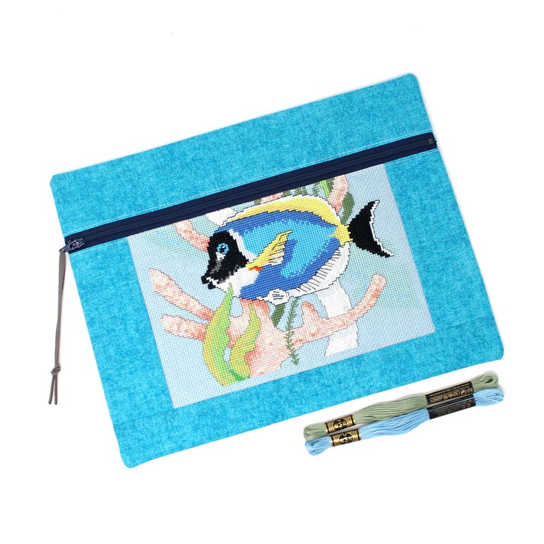 May include: A turquoise needlepoint project bag with a zipper closure. The bag features a cross-stitched design of a blue and yellow fish with black accents, coral, and seaweed. Two skeins of embroidery floss are included.