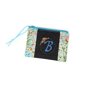 May include: A rectangular zippered pouch featuring a light blue floral pattern on the sides and a black center panel. The black panel displays a cross-stitched letter B in blue, accented with small pink and green floral details and a blue zipper pull.