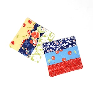 May include: Two quilted fabric coasters with a patchwork design. One coaster features a yellow background with red cherries, a blue background with white flowers, a green background with white flowers, and a navy blue background with red roses. The other coaster features a navy blue background with white flowers, a light blue background with red cherries, and a red background with white polka dots.