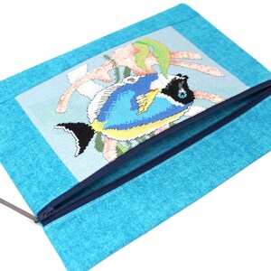 May include: A turquoise zippered pouch featuring a cross-stitched design of a blue and yellow fish with black accents. The fish is set against a light blue background with coral-like elements. The pouch has a dark blue zipper and a gray cord.