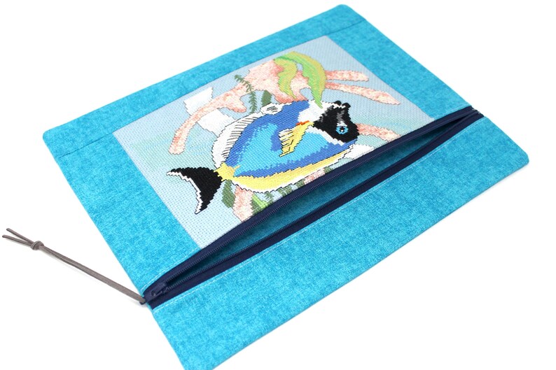 May include: A turquoise zippered pouch featuring a cross-stitched design of a blue and yellow fish with black accents. The fish is set against a light blue background with coral-like elements. The pouch has a dark blue zipper and a gray cord.