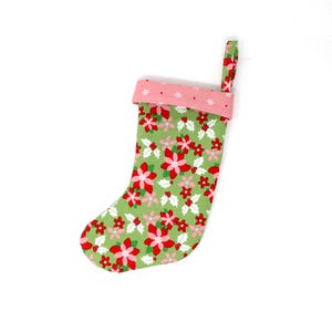 May include: A festive Christmas stocking featuring a green floral pattern with red and pink poinsettias and holly. The cuff is pink with white snowflakes, and a matching loop is attached for hanging.