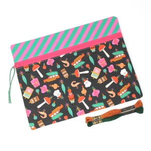 May include: A rectangular black pouch with a pink zip and a green and pink striped top. The pouch is decorated with Christmas-themed illustrations. Two spools of embroidery thread in orange and green are next to the pouch.