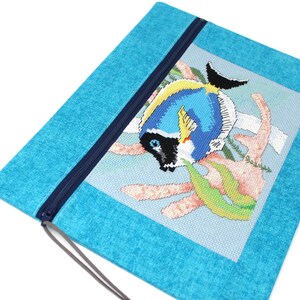 May include: A turquoise zippered pouch featuring a cross-stitched design of a blue, yellow, and black fish with coral and seaweed. The pouch has a dark blue zipper and a matching pull cord.
