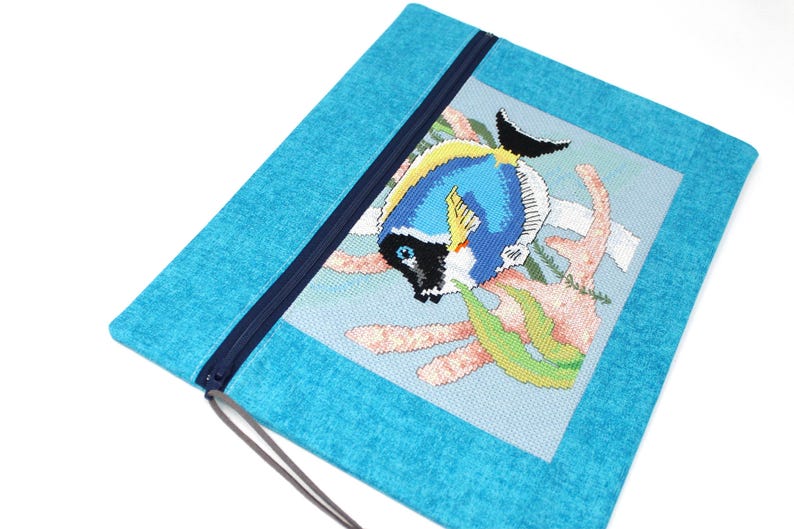 May include: A turquoise zippered pouch featuring a cross-stitched design of a blue, yellow, and black fish with coral and seaweed. The pouch has a dark blue zipper and a matching pull cord.