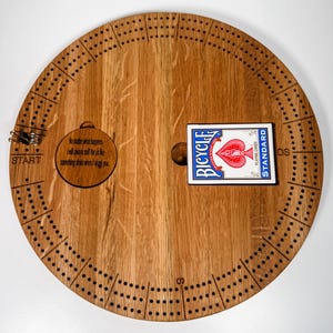 May include: A wooden cribbage board with a deck of Bicycle playing cards. The board has a circular shape with a hole in the center for the cards. The board is made of light brown wood with a dark brown stain. The board has a quote engraved on it: "No matter what happens I will always sniff the air like something stinks when I snag you."
