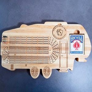 May include: Wooden cribbage board shaped like a camper with a deck of Bicycle playing cards and a small wooden circle with the text "It's okay if you don't like cribbage. It's kind of a weird people game anyway."