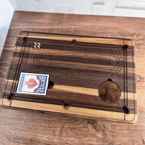 May include: A wooden cribbage board with a playing card in the card holder. The board is made of walnut and maple wood and has a dark brown stain. The board has a track for the pegs and a scoring area. The board also has a small circle with the text "Charlie on the cage" and a pair of crossed hockey sticks.