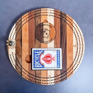 May include: A wooden cribbage board with a circular playing surface and a card holder. The board is made of different types of wood, creating a striped pattern. The board has a circular inscription that reads "Shake a peg it's cribbage time!" and a playing card is in the card holder.