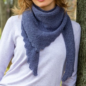 May include: A knitted, blue-grey scarf with a scalloped edge. The scarf is draped around the neck and shoulders. The person in the image is wearing a light purple sweater. The background is blurred, with a tree trunk visible on the right.