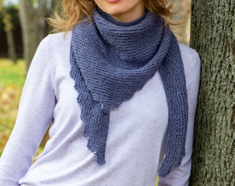 May include: A knitted, blue-grey scarf with a scalloped edge. The scarf is draped around the neck and shoulders. The person in the image is wearing a light purple sweater. The background is blurred, with a tree trunk visible on the right.