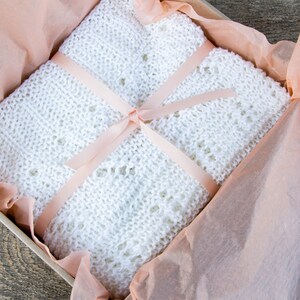May include: A white knitted baby blanket with a delicate pattern is tied with a peach-colored ribbon and placed in a gift box lined with tissue paper.