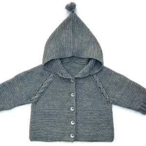 May include: A gray knitted baby cardigan with a hood and pom-pom detail. The button-down sweater features a textured knit design, long sleeves, and decorative stitching along the shoulders. The hooded sweater is a cozy garment for infants.